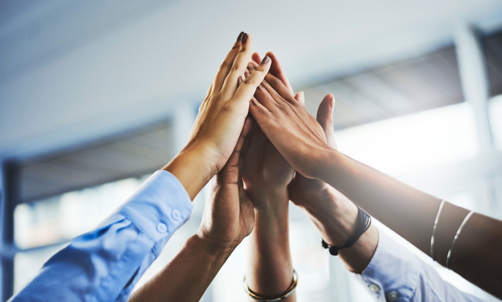 A low-angle shot of several hands, belonging to a diverse group of people, stacked on top of each other in a high-five gesture, symbolizing teamwork and unity. The background is blurred, showing an indoor office setting.