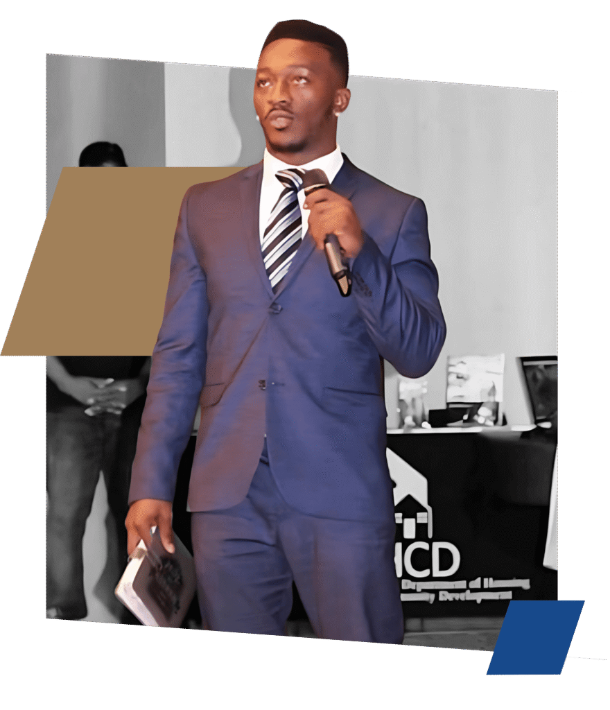 A man in a blue suit and striped tie holds a microphone and a book while speaking at an event. A logo for the Department of Housing and Community Development is visible in the background.