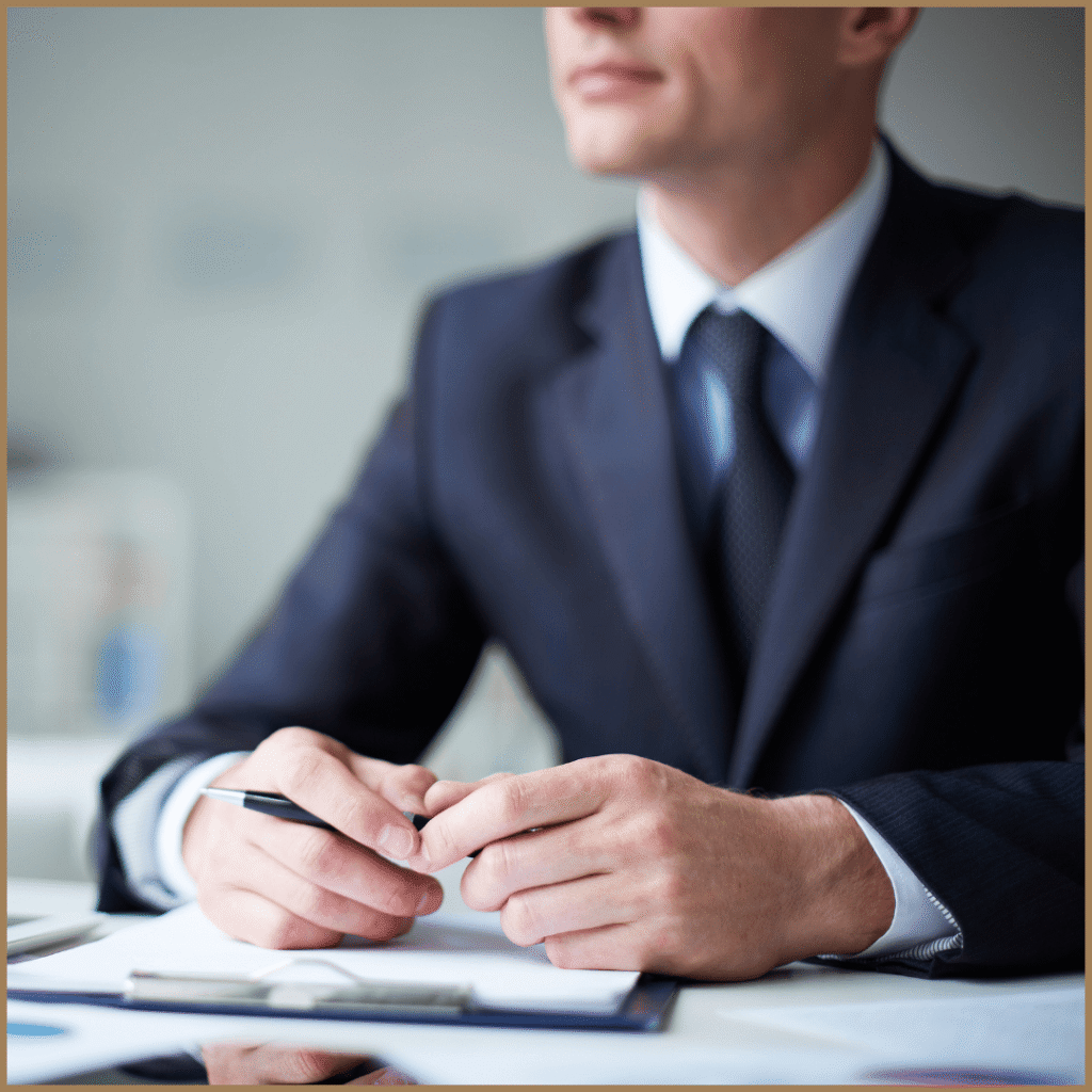 A close-up, cropped photo of a person in a dark suit and tie sitting at a desk. Their hands are clasped, and they are holding a pen.