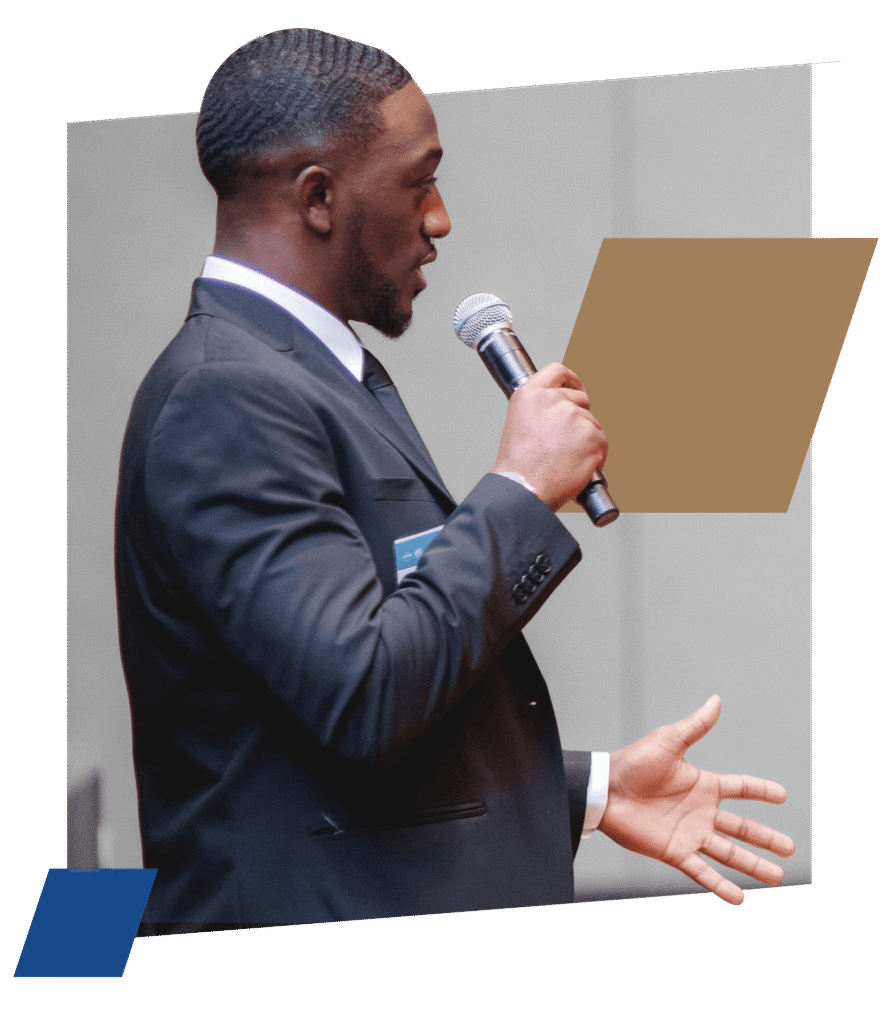 A man in a dark suit speaks passionately into a microphone while gesturing with his hands. The background has a gray wall with abstract brown and blue shapes.