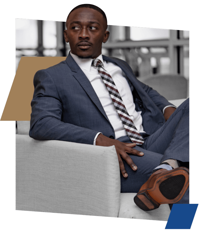 A man in a blue suit and a plaid tie is seated in a gray chair, with one leg crossed over the other, and is looking off to the side.