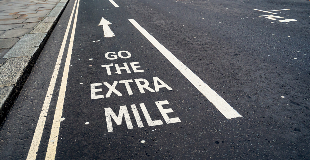 A close-up view of asphalt road with white painted markings. The phrase "GO THE EXTRA MILE" is prominently painted in large white capital letters with an arrow pointing forward, next to double yellow lines and a solid white line.