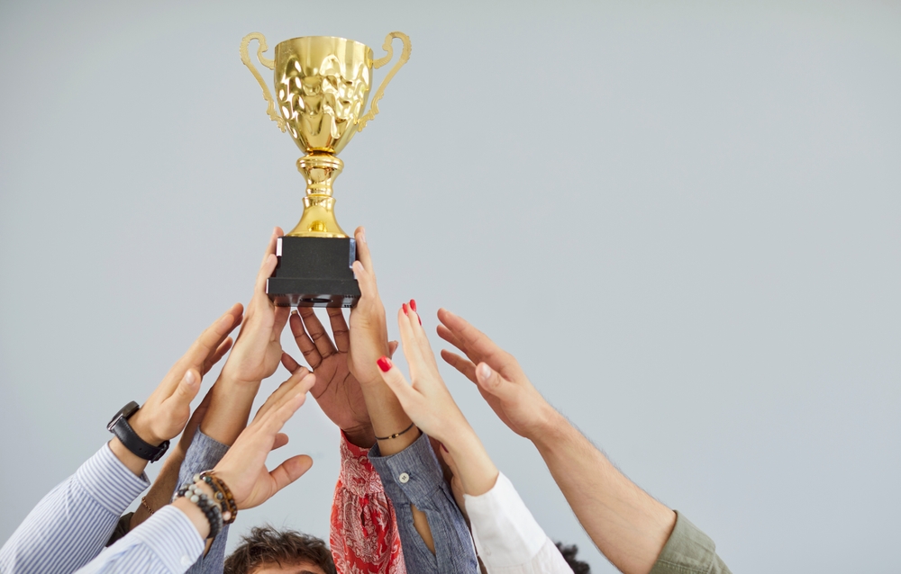 Multiple hands from a diverse group of people are raised upwards, collectively supporting and reaching towards a gleaming golden trophy with a black base, set against a plain light gray background.