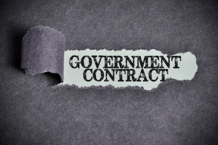 A piece of gray paper has been torn open to reveal the words "GOVERNMENT CONTRACT" in bold, black lettering on a white background. The torn edge of the paper is curled back to the left.