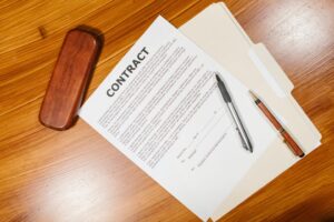 A high-angle shot of a contract document placed on a wooden desk. A brown wooden box, a silver pen, and a brown pen are placed beside the contract, which is resting on top of a manila folder.