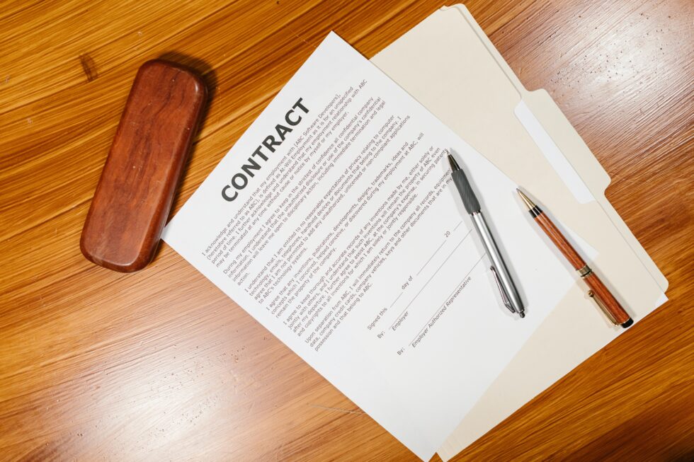 A high-angle shot of a contract document placed on a wooden desk. A brown wooden box, a silver pen, and a brown pen are placed beside the contract, which is resting on top of a manila folder.