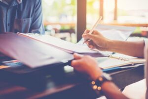 A close-up shot of a person's hands signing a document on a clipboard with a pen. The person is sitting at a table across from another individual, visible in the upper left, with warm sunlight coming through a window in the background.