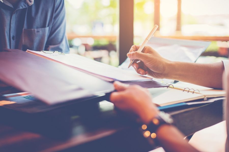 A close-up shot of a person's hands signing a document on a clipboard with a pen. The person is sitting at a table across from another individual, visible in the upper left, with warm sunlight coming through a window in the background.