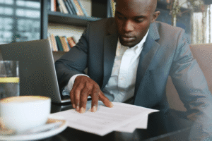 A Black man in a dark gray suit and white shirt sits at a desk, intently reading a document. A laptop, a glass of water, and a coffee cup are also on the desk. A bookshelf is in the blurred background.